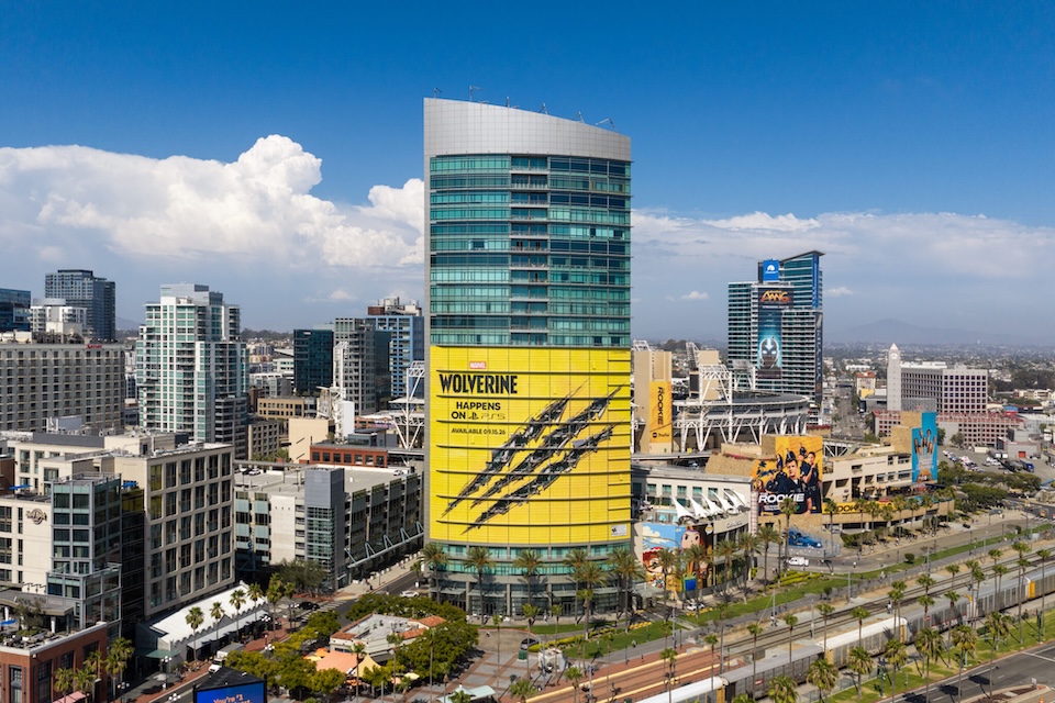 default | Muse by Clios Aerial cityscape with tall glass buildings and a large yellow Wolverine billboard on a curved tower in the foreground.