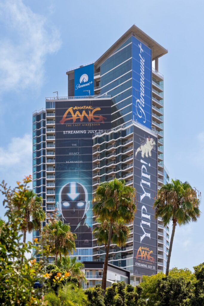 KAP_CC26_Install_Park12_Paramount_High_Res-1-scaled | Muse by Clios Tall modern hotel/condo tower adorned with large Avatar: The Last Airbender banners and a Paramount+ sign against a blue sky.
