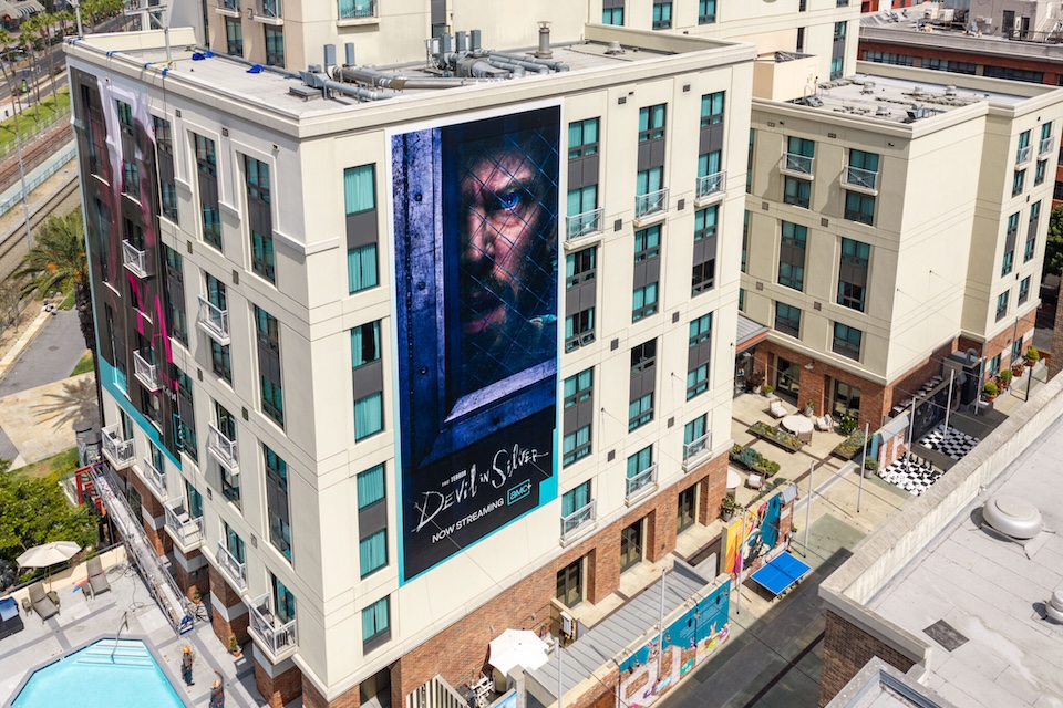default | Muse by Clios Tall beige apartment building with a large vertical banner showing a man's face and the words 'Devil in Silver' advertising now streaming/Netflix-like service.