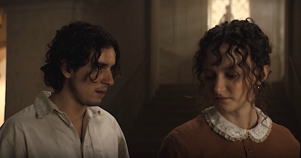 Two young adults stand in a dim hallway, facing each other and sharing a quiet moment. She wears a brown dress with a lace collar; he in a light shirt.