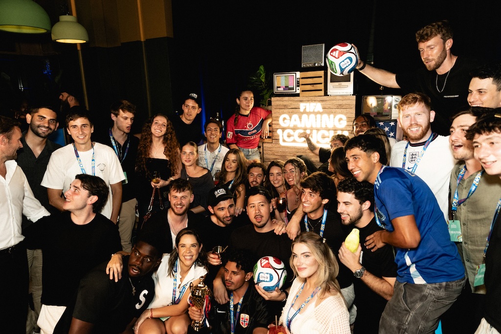 Miami_1 | Muse by Clios Group of diverse young adults posing for a photo at a FIFA Gaming Lounge event, many wearing lanyards and smiling, with a soccer ball and trophy visible in front.