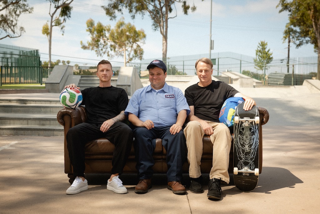 Tony Hawk x Marco Reus_2 | Muse by Clios Three men sit on a brown leather sofa outdoors at a skatepark; left holds a soccer ball, middle wears a uniform, right rests a skateboard with helmet nearby.
