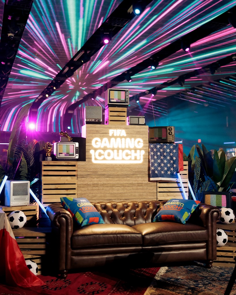 Couch | Muse by Clios Retro gaming lounge with CRT TVs on wooden pallets, a neon-lit sign reading 'FIFA GAMING COUCH', and a brown leather sofa.