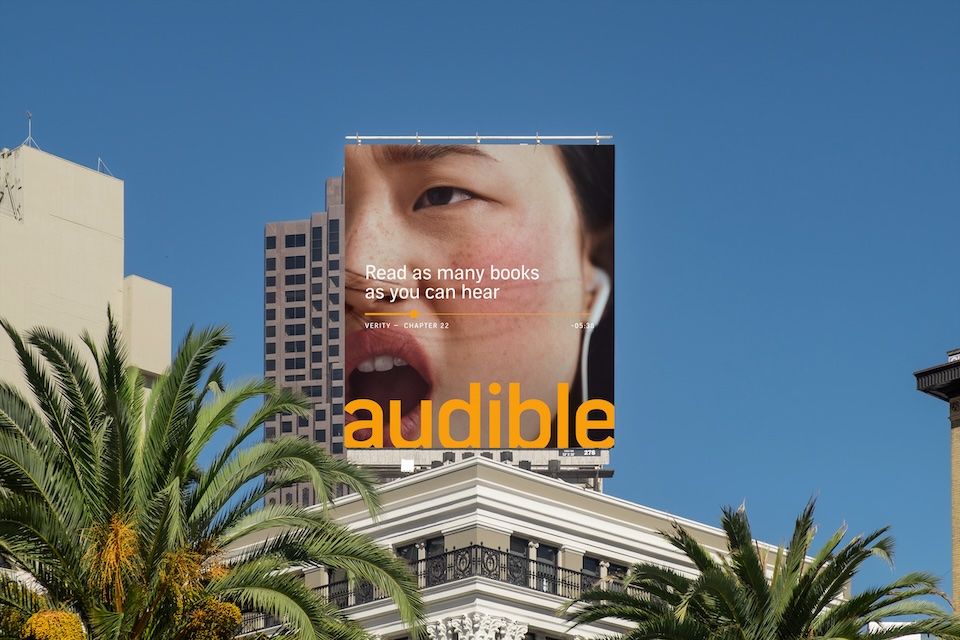 Billboard ad for Audible showing a close-up face and the orange 'audible' logo above a white building with palm trees and a blue sky.