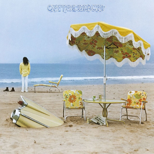 Beach scene with a yellow umbrella, floral chairs, a table, and surfboards in the sand; person in a yellow jacket faces the ocean.