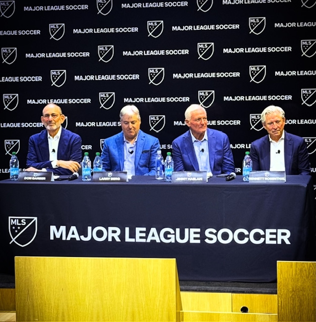 Four men in blue jackets sit at a press conference table with MLS Major League Soccer logos on a black backdrop and table banner.