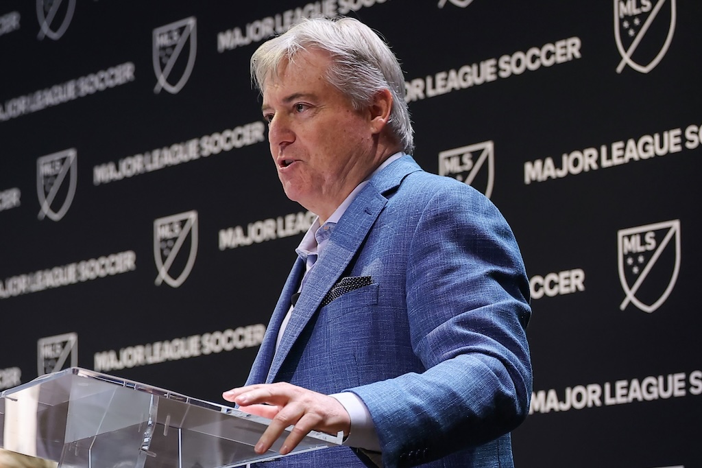 Man in a blue suit speaks at a clear podium during a Major League Soccer event backdrop says MLS and major league soccer
