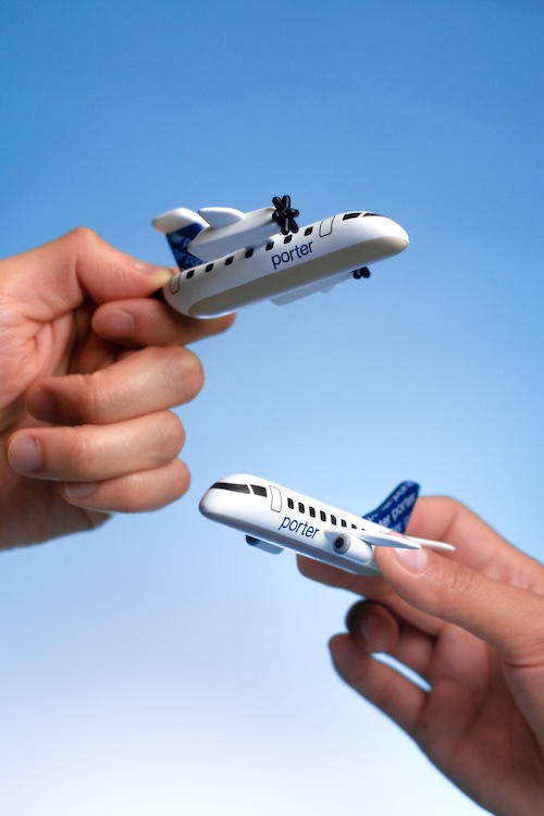 porter 2 | Muse by Clios Two small Porter toy airplanes held between fingers against a blue sky background.