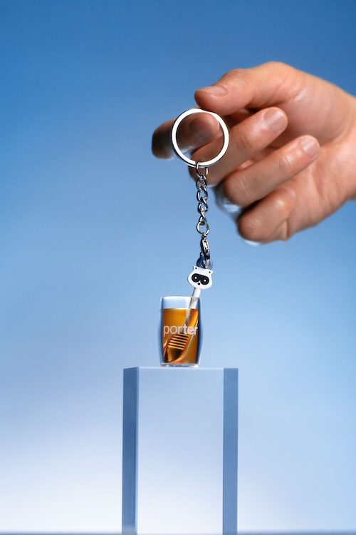 porter 3 | Muse by Clios Hand holding a keychain with a miniature Porter beer bottle charm balanced on two blue pillars, against a blue sky.