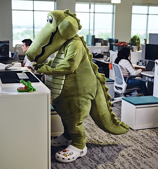 Person in a green dinosaur costume typing at a desk in a bright office environment.