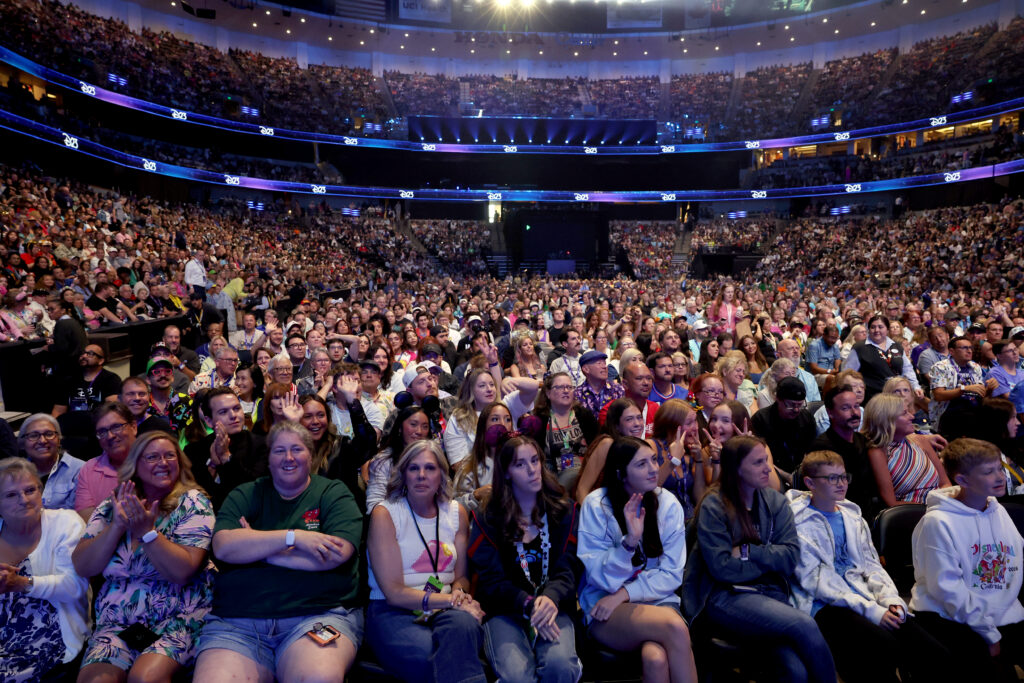 Large crowd seated in an arena, facing a stage with blue lights and screen backdrop.