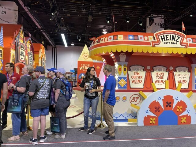 Bright Heinz expo booth with carnival-style arches and a large wheel display; attendees line up while two staffers chat near the center of the booth.