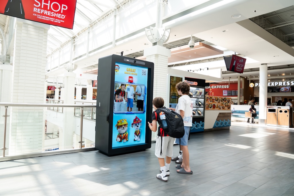Two children with backpacks examine a tall digital kiosk in a bright shopping mall.