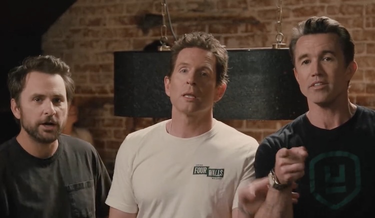 Three men stand in a brick-walled studio, facing the camera. Center man wears a white T-shirt with a small logo; left and right men wear dark shirts, with the right man pointing toward the lens.