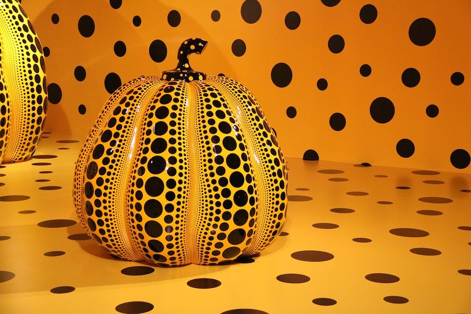 Y2 | Muse by Clios Large orange pumpkin sculpture covered in black polka dots in a yellow gallery room with matching dot patterns on walls and floor.