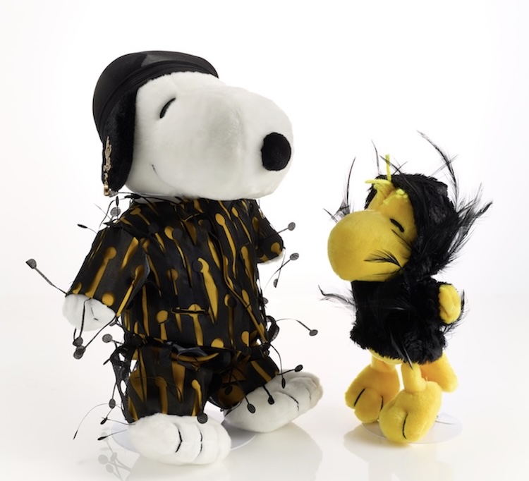 Screenshot | Muse by Clios Snoopy plush in a black and yellow patterned outfit with hanging bead accessories, facing Woodstock plush in a black feathered costume.