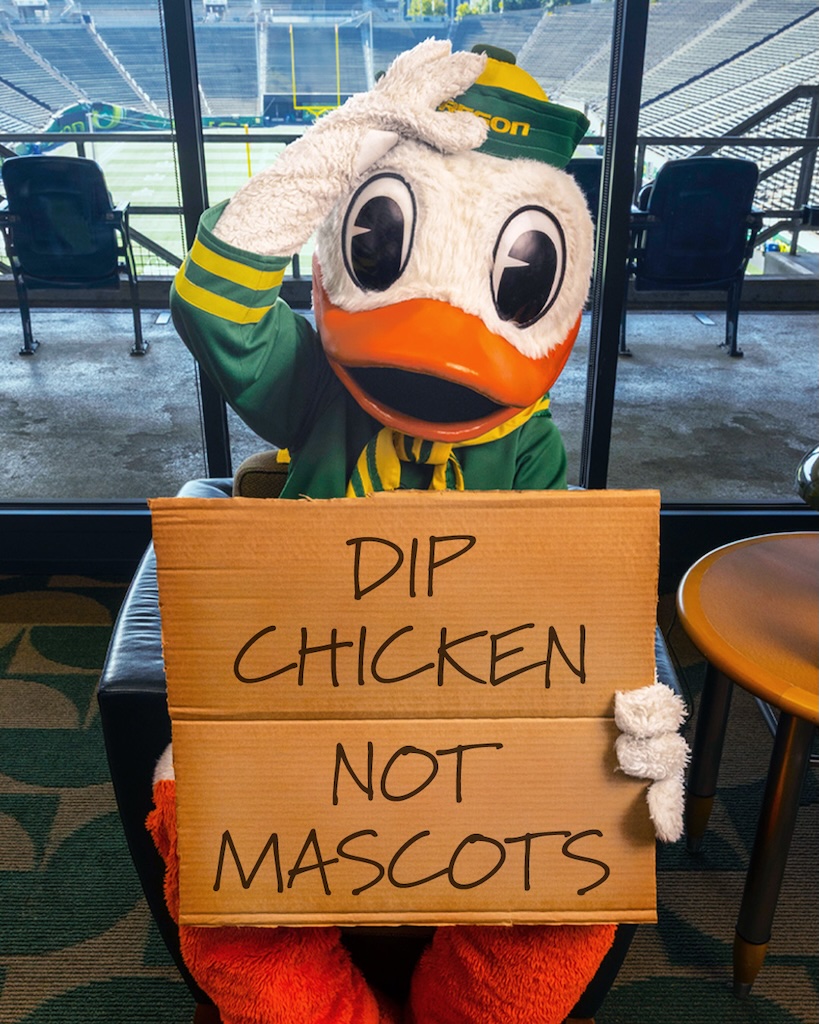 Duck mascot in green and yellow outfit holds a cardboard sign that reads 'DIP CHICKEN NOT MASCOTS' in a stadium seating area.