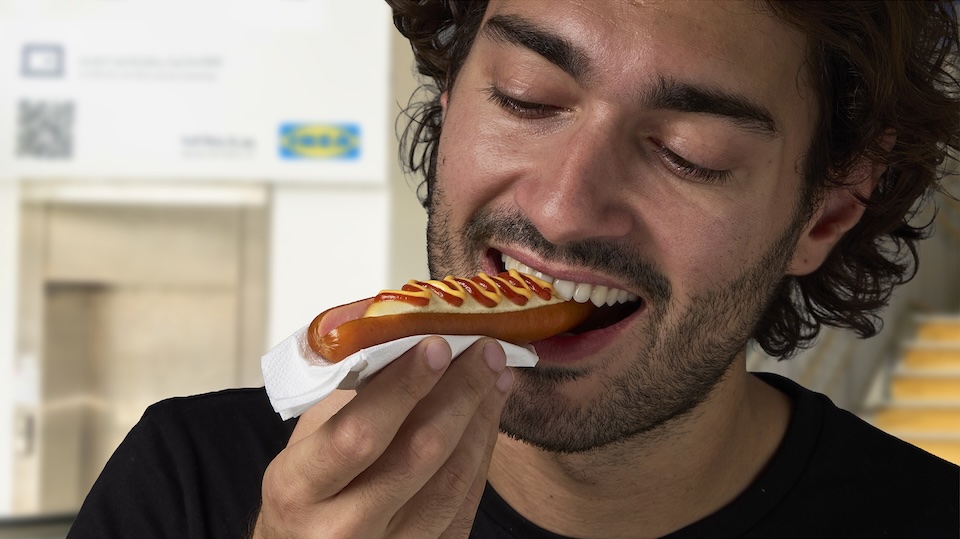 Close-up of a man with dark hair and stubble biting into a hot dog topped with mustard, held in a napkin to catch drips.