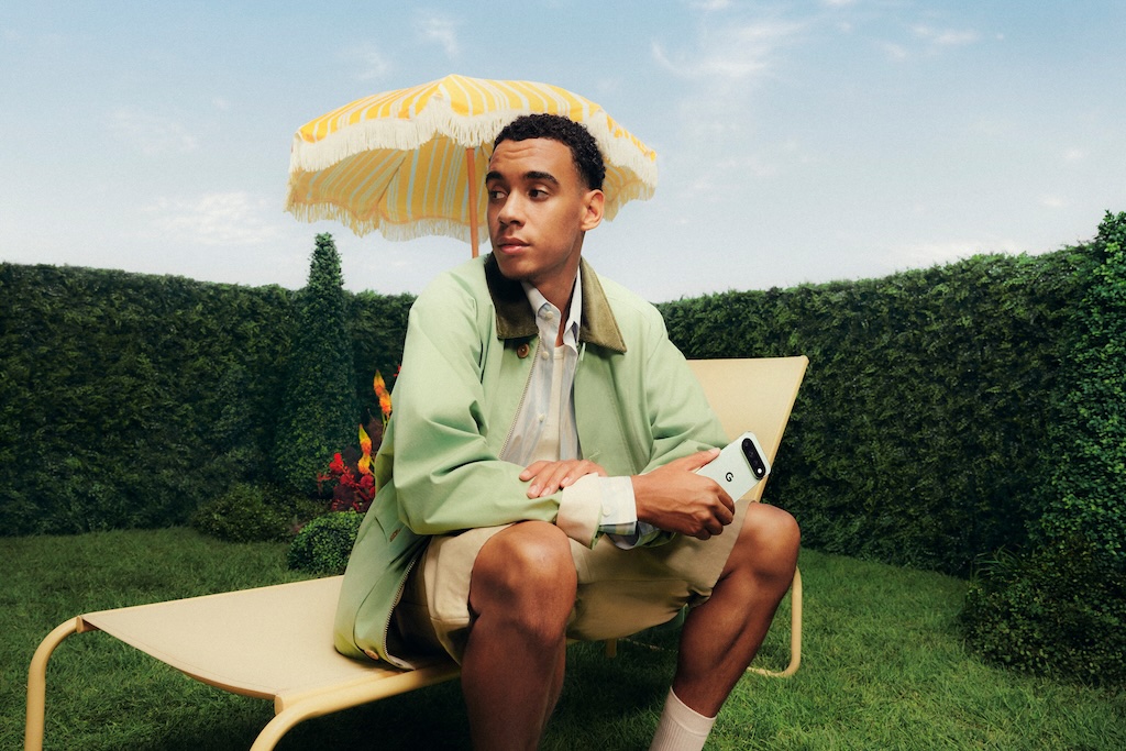 AKQA_GooglePixel-JamalMusiala_05 | Muse by Clios Young man sits on a pale yellow outdoor lounge chair under a striped yellow umbrella in a garden, holding a white smartphone.