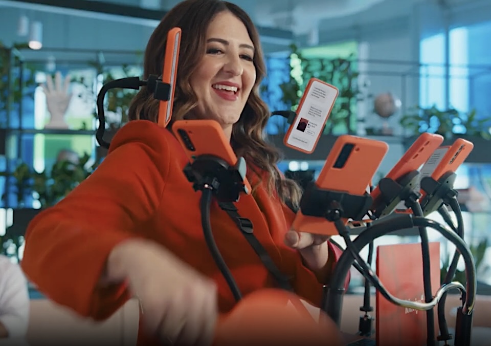 Screenshot | Muse by Clios Smiling woman in a red jacket demonstrates multiple orange smartphones mounted on adjustable stands in a bright tech workspace.