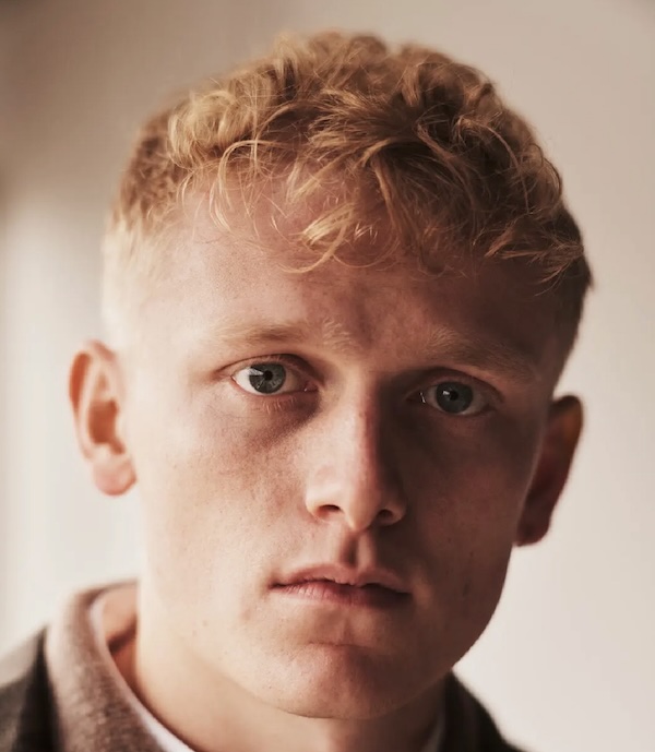 Close-up portrait of a young man with short blond hair looking directly at the camera, neutral expression.