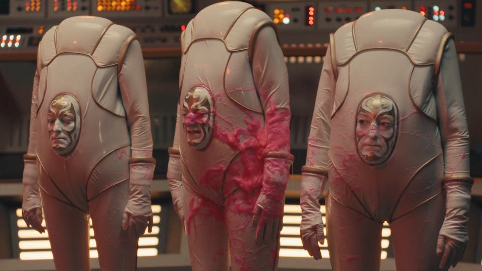 Three people in beige sci‑fi suits stand in a high‑tech control room; pink stains smear their suits and faces through circular chest windows.