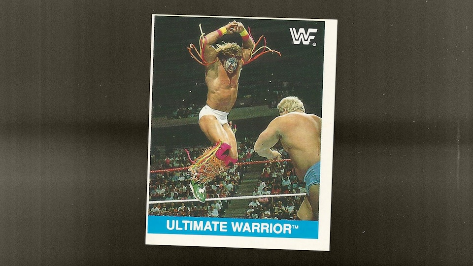 WWE trading card showing Ultimate Warrior leaping in the air with arms raised during a wrestling match, audience in the background and the blue 'Ultimate Warrior' banner at the bottom