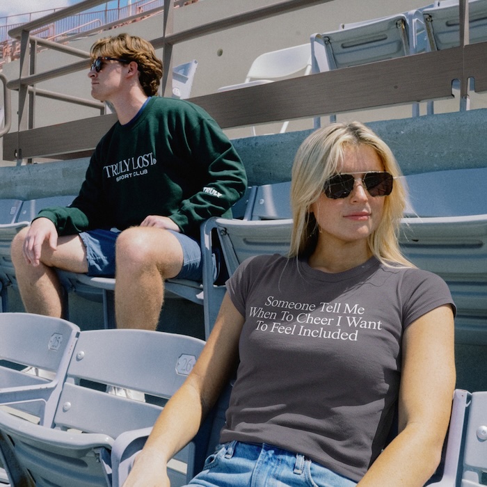 Two people sit in stadium bleachers; the woman in front wears sunglasses and a gray T-shirt that reads,'Someone Tell Me When To Cheer I Want To Feel Included'.