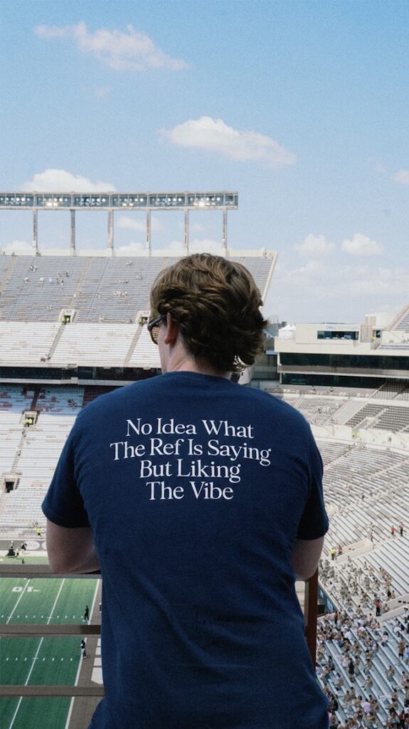 Spectator facing away in a stadium stands wearing a blue T-shirt that reads: “No Idea What The Ref Is Saying But Liking The Vibe.” and a football field visible below.