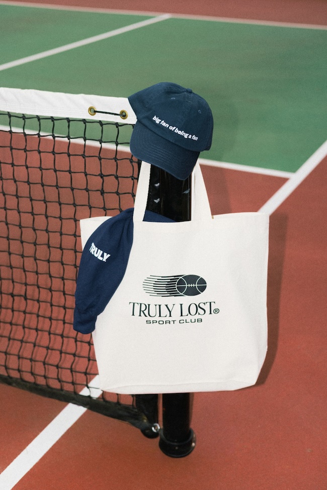 White tote bag with the'Truly Lost Sport Club' logo hanging on a tennis net, with a navy cap resting on top above the bag on a tennis court edge.