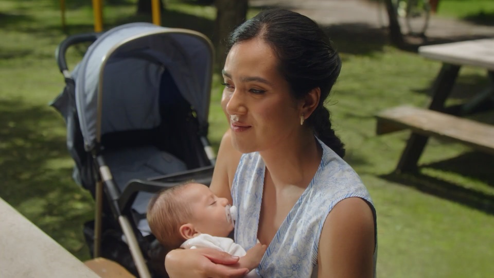 Mother holding a sleeping baby in her arms at a sunny park, with a stroller in the background.
