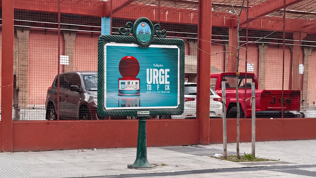 Tulipán_Urge to f ck_OOH evidence 1 | Muse by Clios Outdoor advertisement board for perfume on a green pedestal, with a blue bottle and red cap, behind a fence and parked cars.