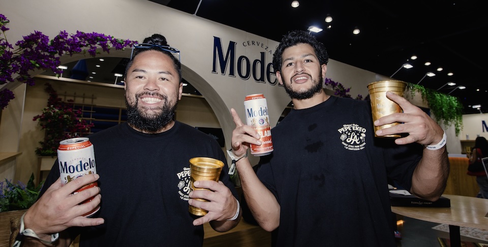 Two men with beards at a Modelo booth, each holding a beer can and a gold cup, smiling at the camera.
