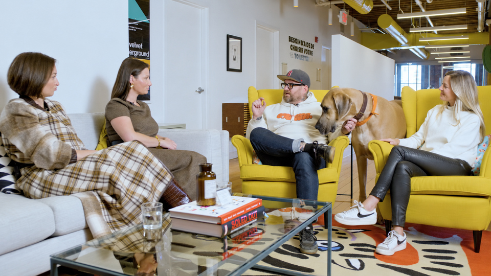 Group of four adults seated in a modern lounge talking, with a large dog standing by a man in a cap beside a yellow armchair.