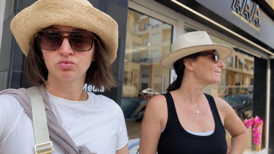 Two women wearing sun hats and sunglasses posing outside a storefront. The foreground woman wears a white shirt and beige bag strap; one wears a black tank top in the background.