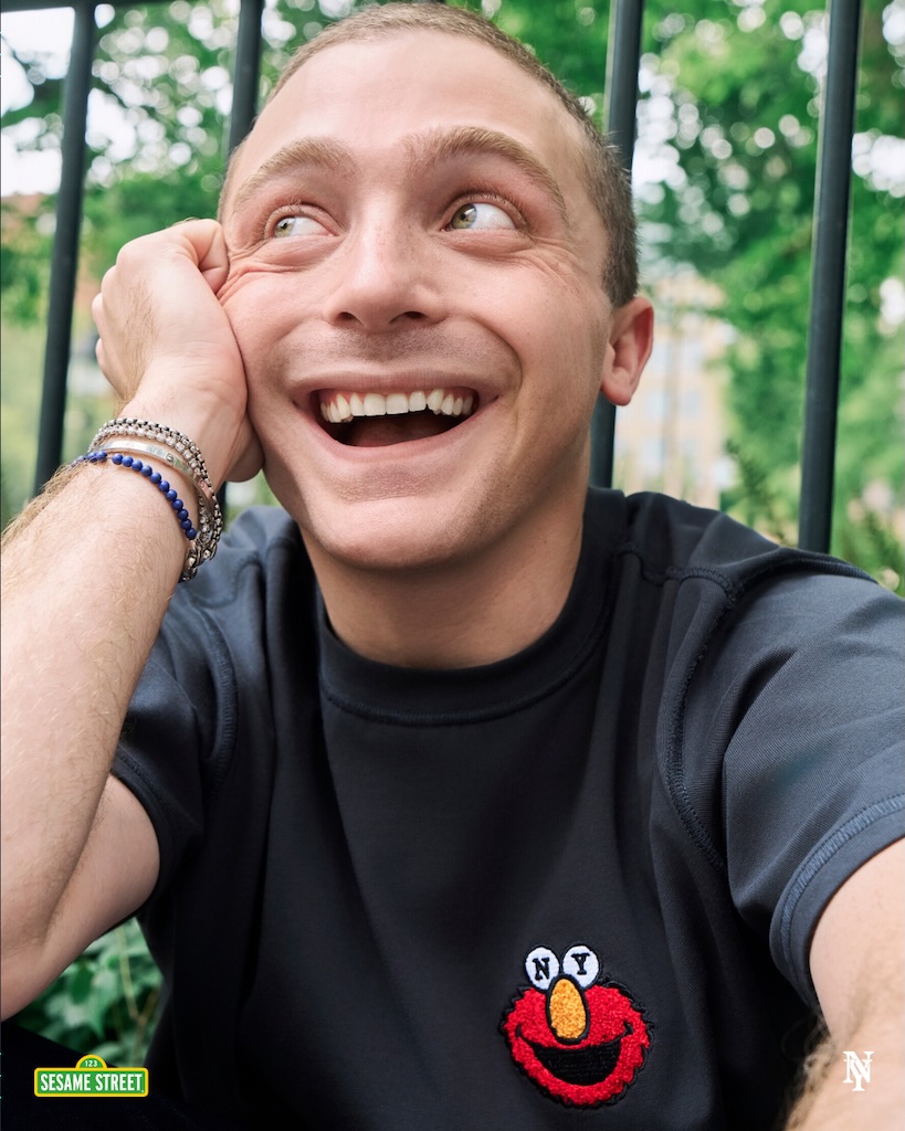 Jake Shane | Muse by Clios Young man smiling broadly, resting his cheek on his hand, in a black shirt with a Sesame Street patch outdoors by a fence.