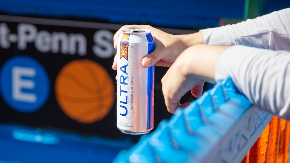 Close-up of a person’s hands holding a silver Ultra energy drink can, with a sports banner in the blurred background.