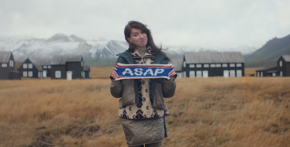 Woman standing in a grassy field holding a blue scarf that reads ASAP, with dark houses and snow-covered mountains in the background.